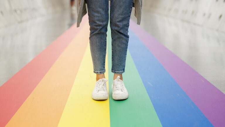 Føtter i joggesko står på regnbuefarget gate, symbol på Pride og mangfold.